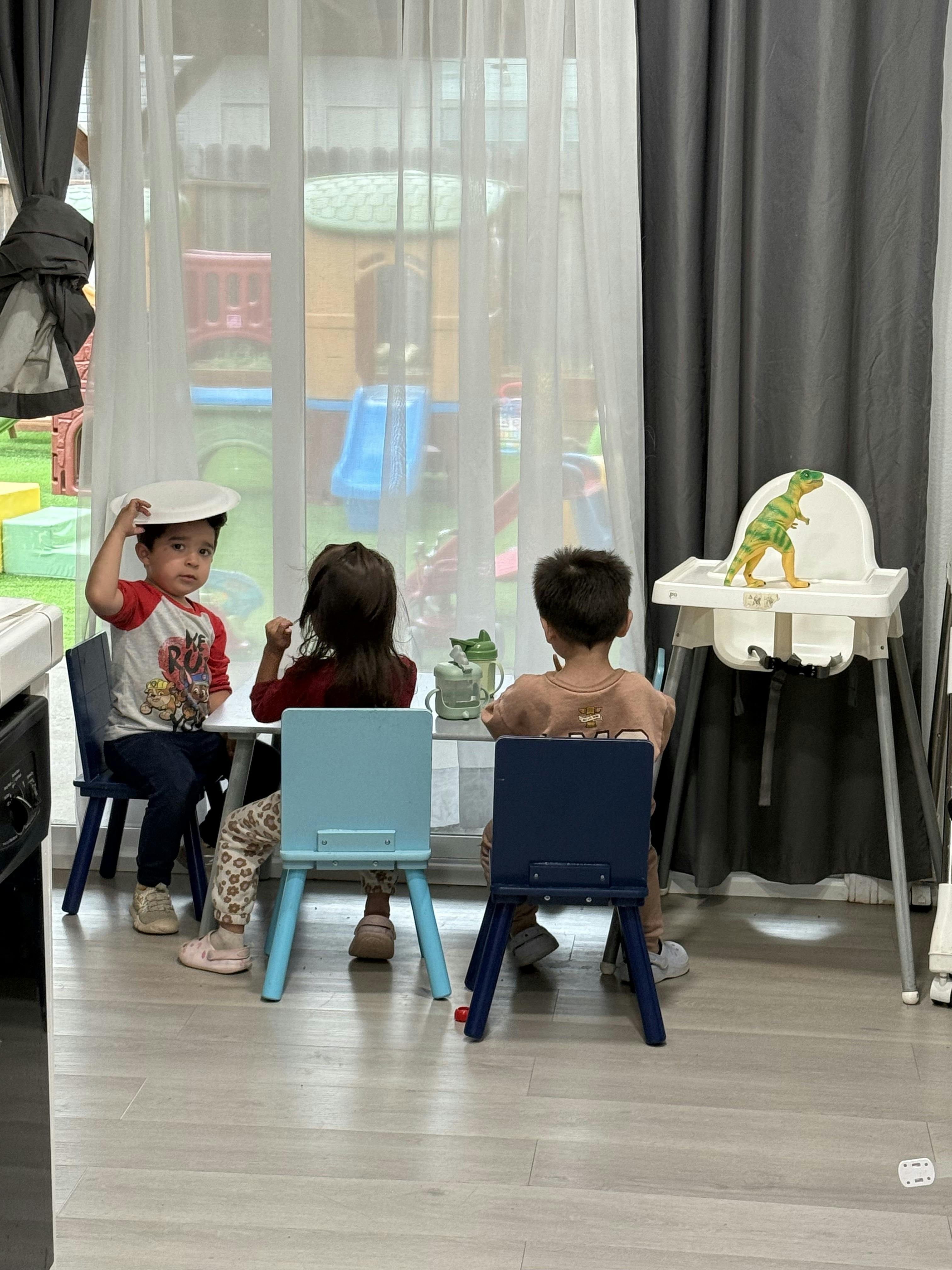 Three children sitting at a small table indoors with a toy dinosaur nearby