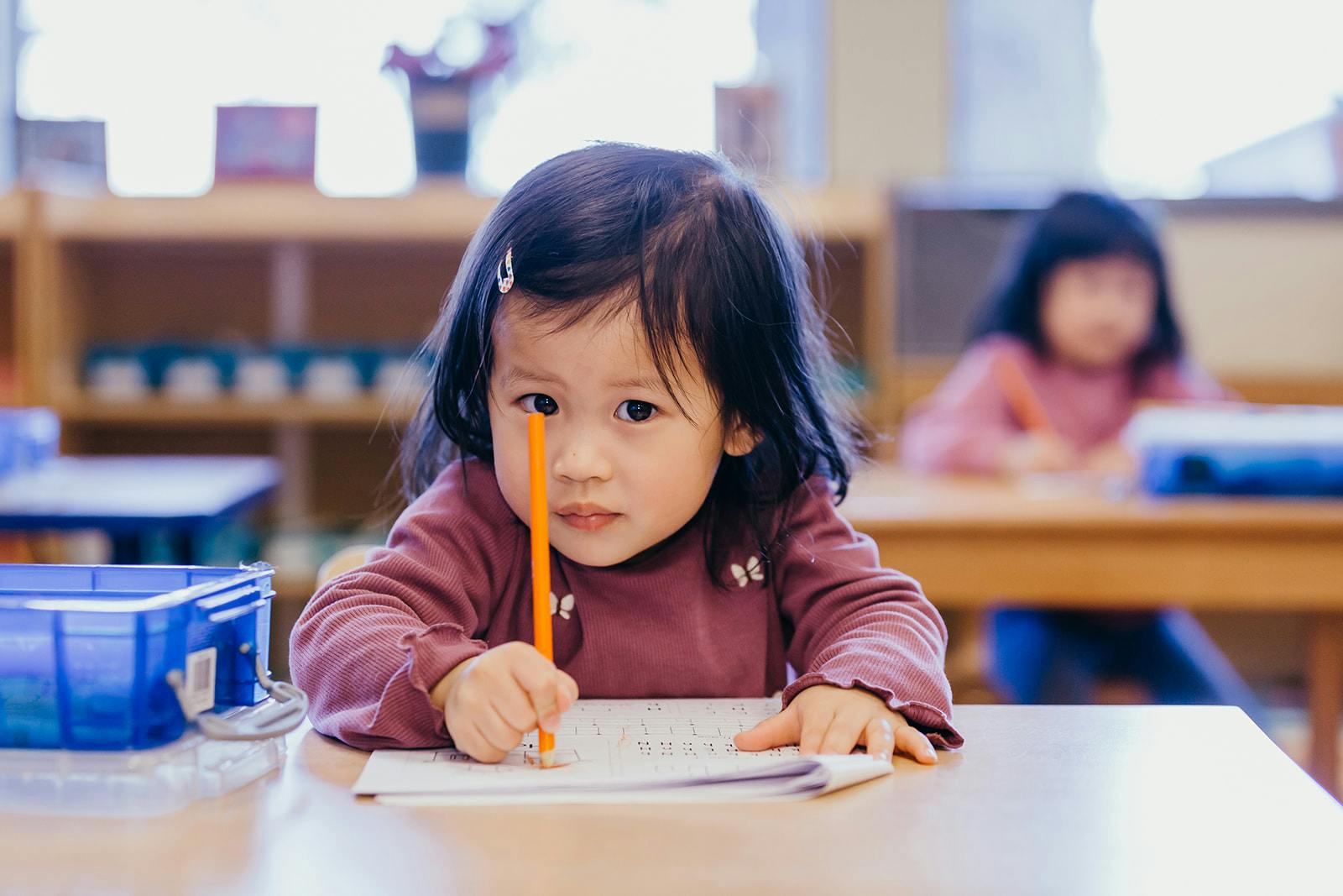 Child holding a pencil over paper at a desk in a classroom