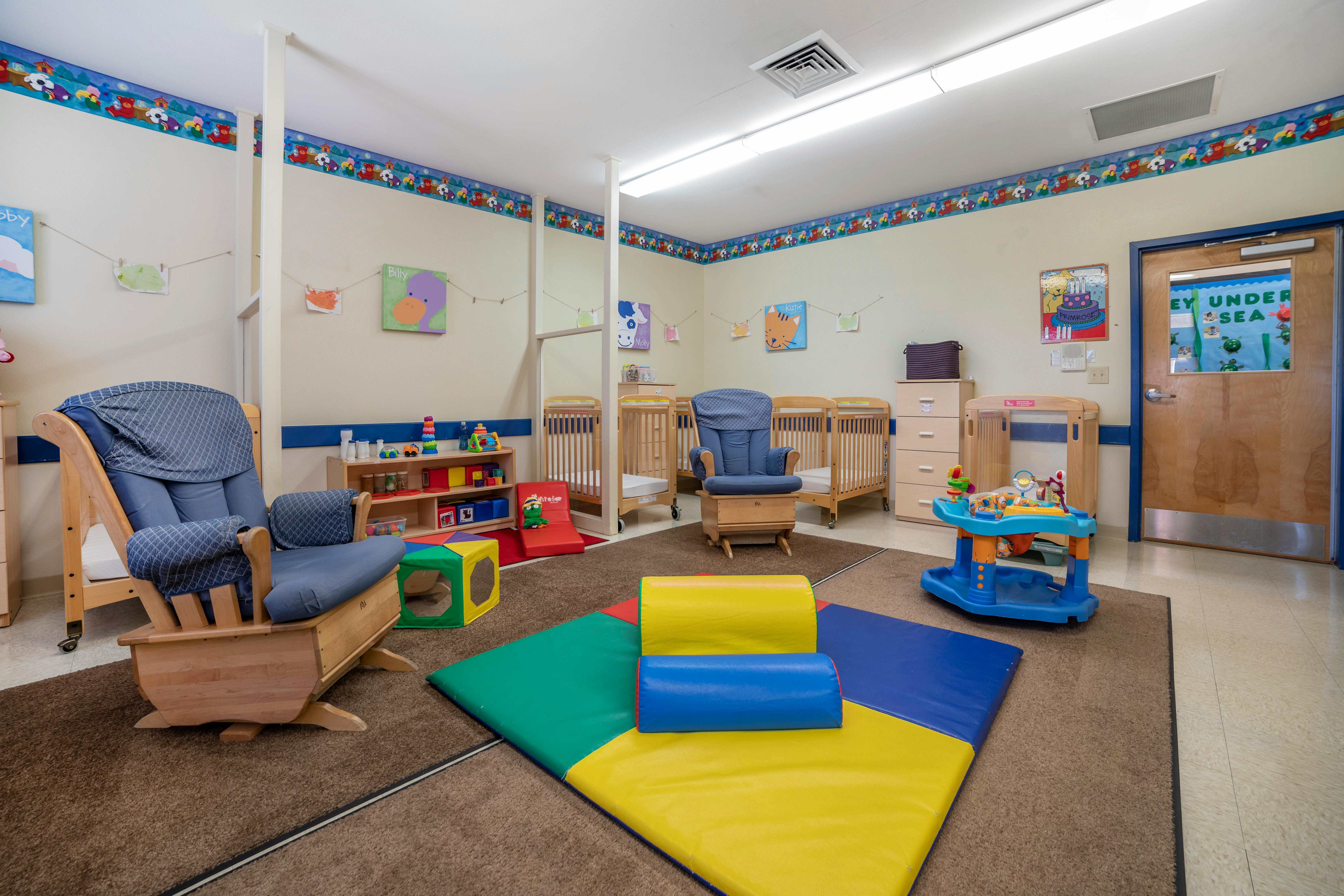 A colorful childcare classroom with cribs, chairs, and play mats