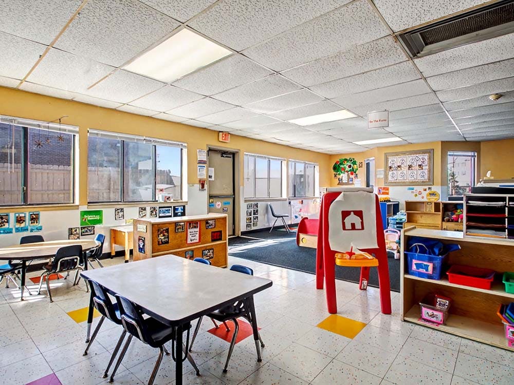 Empty classroom with small tables and chairs, shelves, and art easel