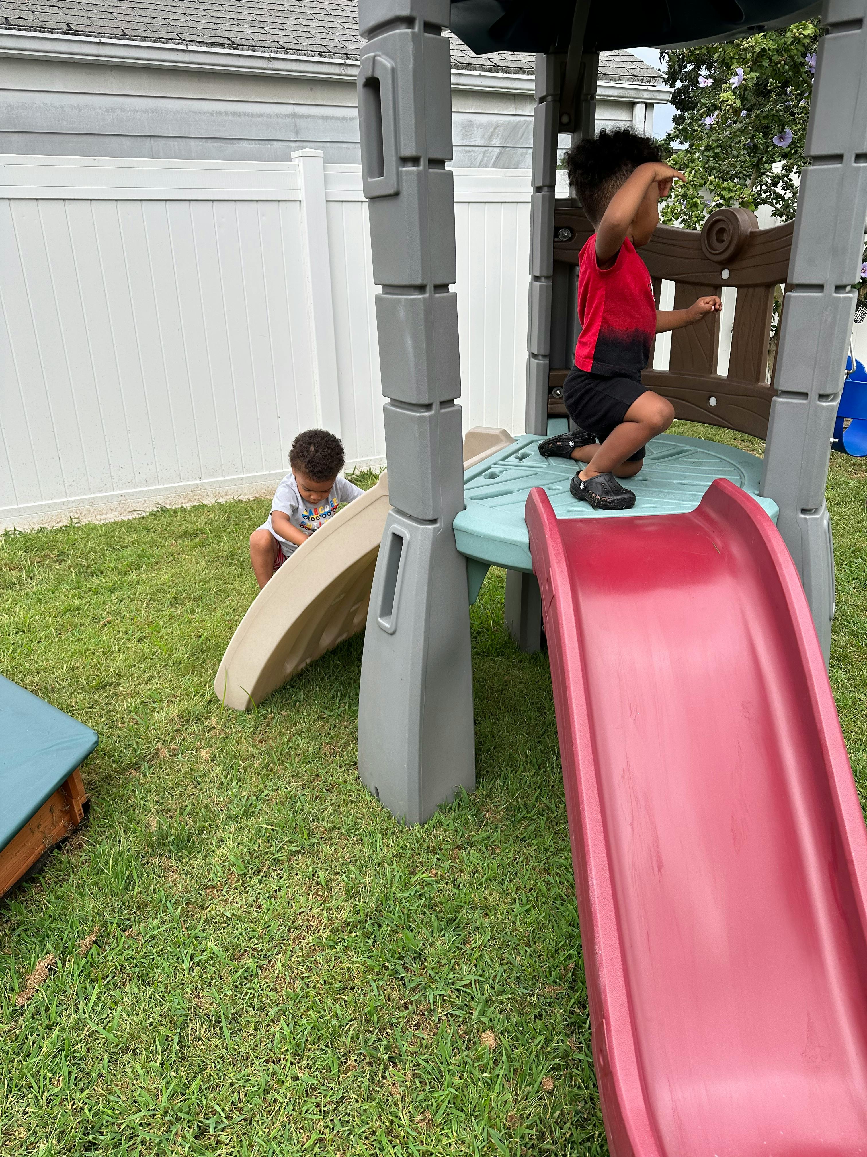 Two children playing on a slide and play structure in a grassy yard