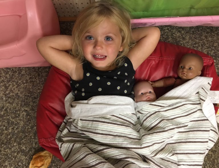 Child lying on a red cushion with two dolls wrapped in blankets
