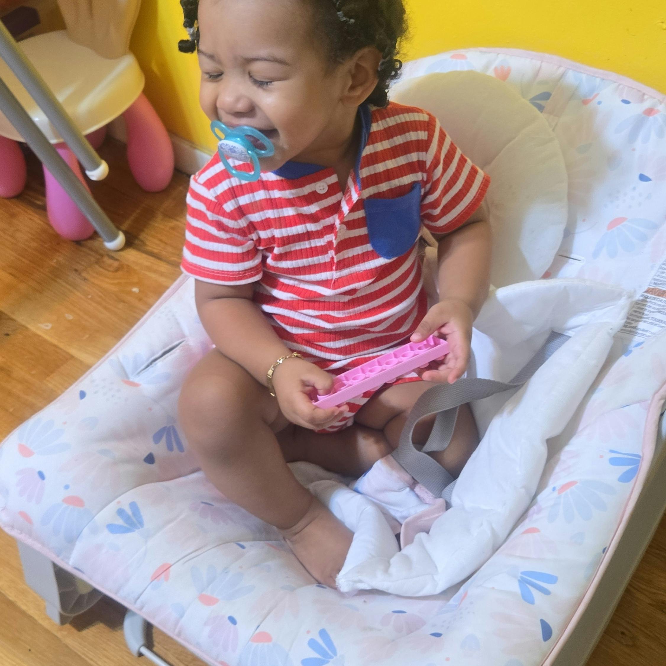 Child in a striped shirt with a pacifier in a baby bouncer holding a toy