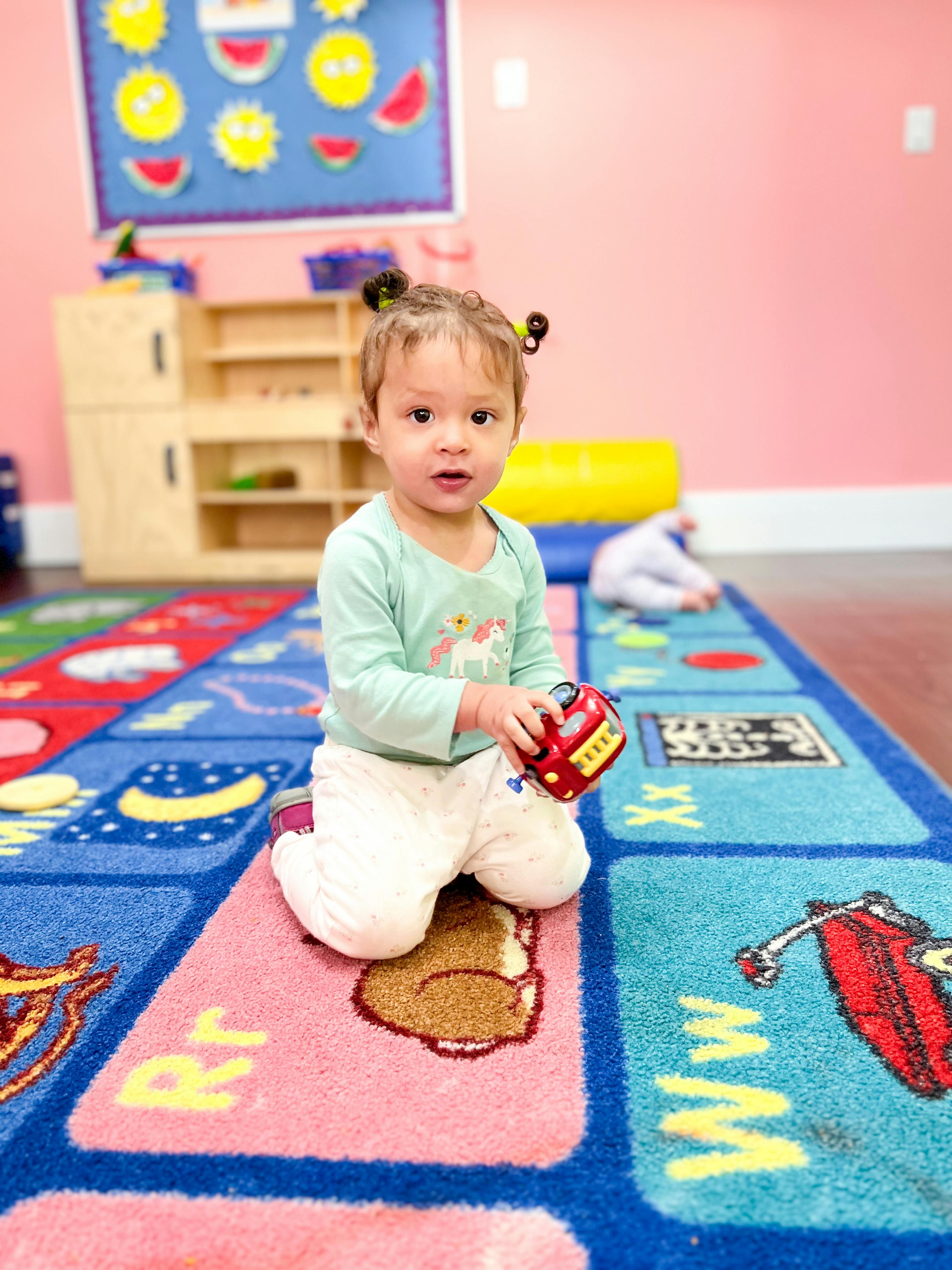 Child kneeling on a colorful rug in a classroom, holding a small red toy