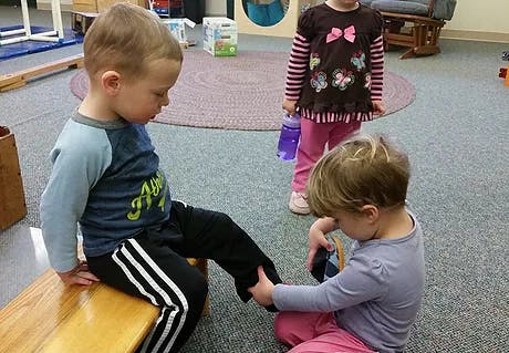 Child sitting on bench while another child helps with their shoe