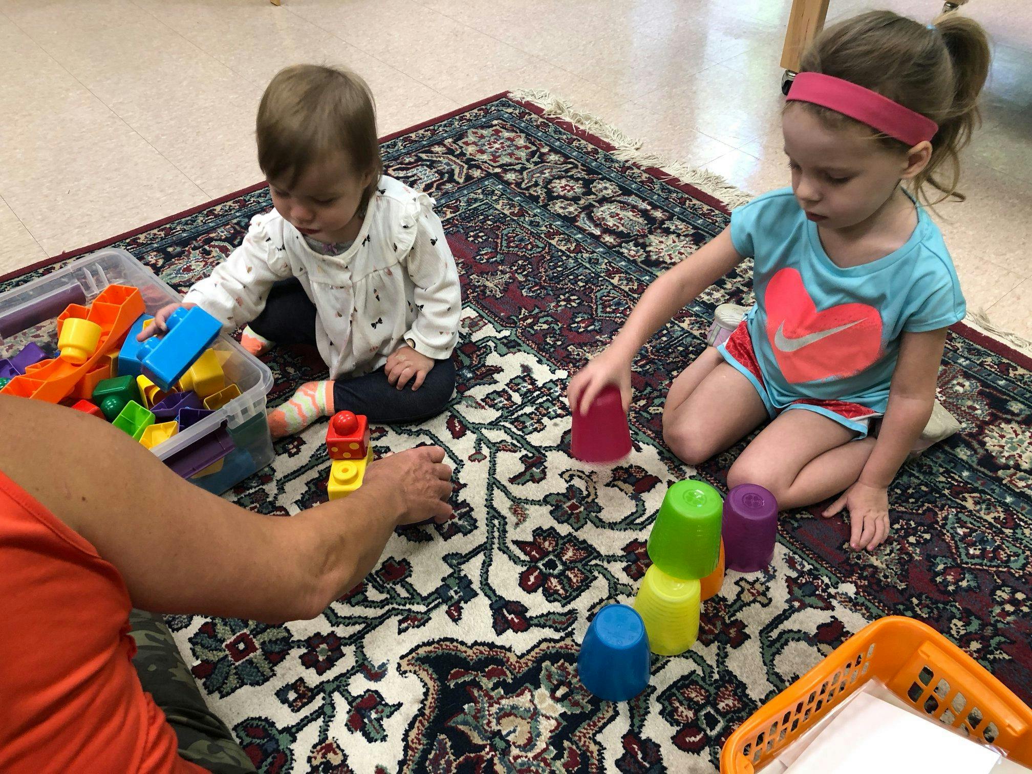 Two children and an adult playing with blocks and cups on a rug