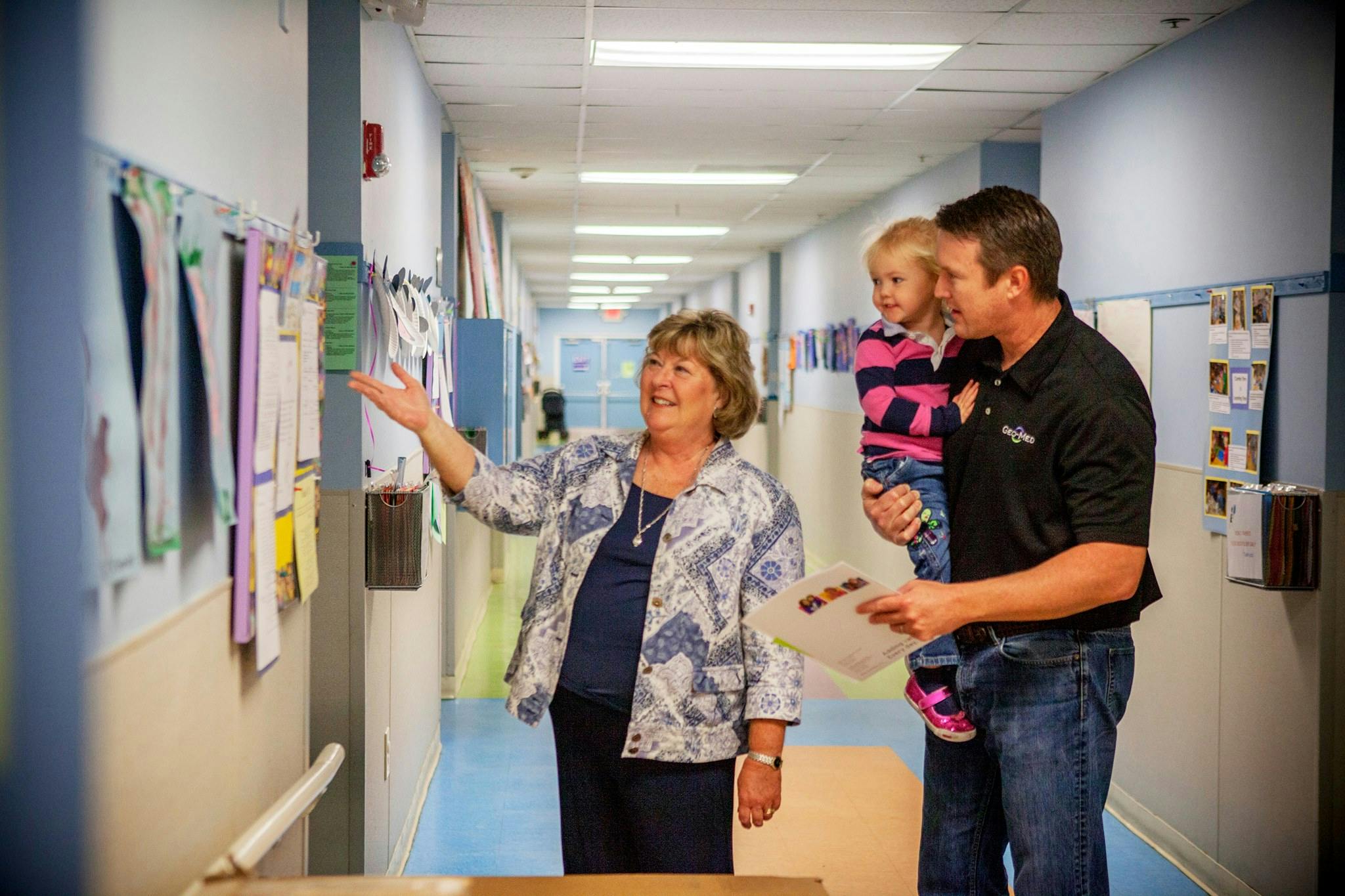 Adult pointing at papers on a wall with an adult holding a child nearby