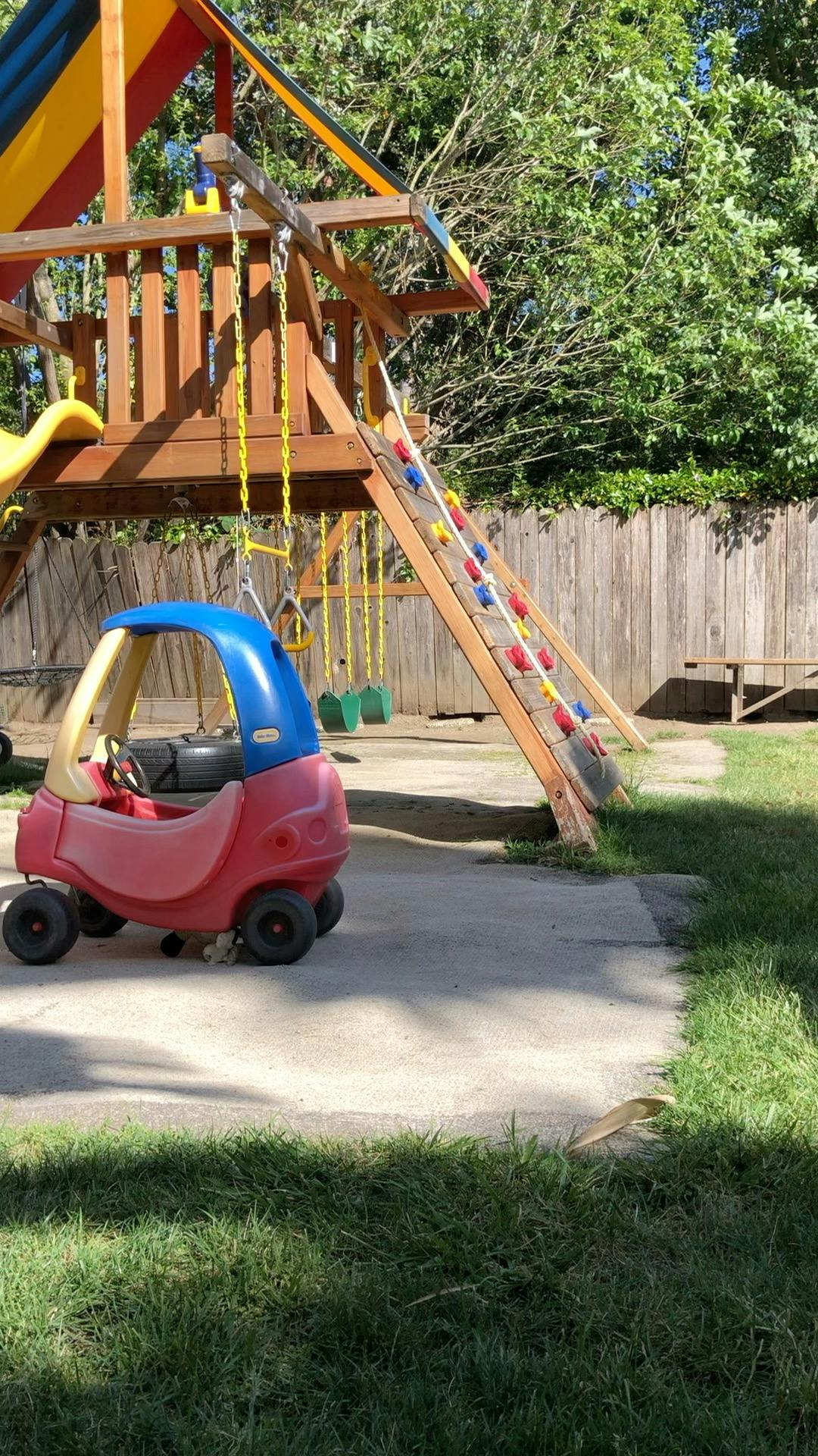 Toy car on a concrete path next to a wooden playset with swings and a slide