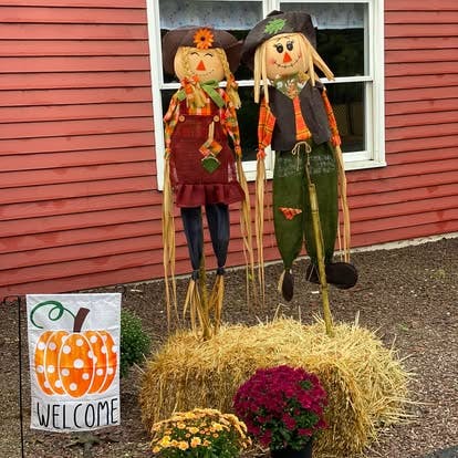 Two scarecrows on hay bales with a welcome sign and potted flowers