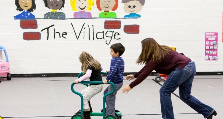 Person helping two children on riding toys in a room with "The Village" on the wall