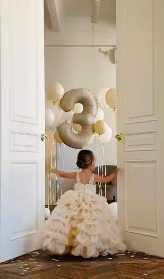 Child in a ruffled dress looking at a large number 3 balloon and other balloons