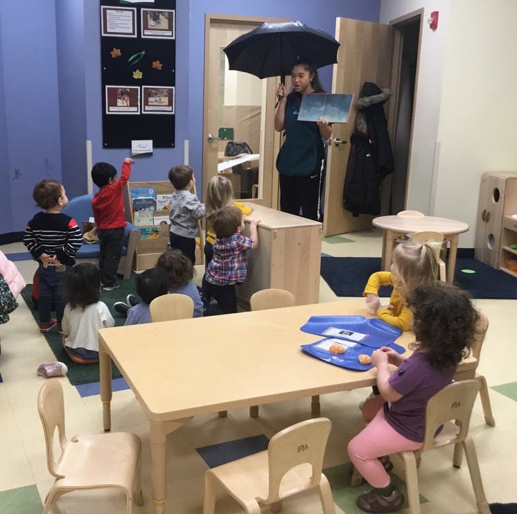 Person holding an umbrella and book, speaking to a group of children