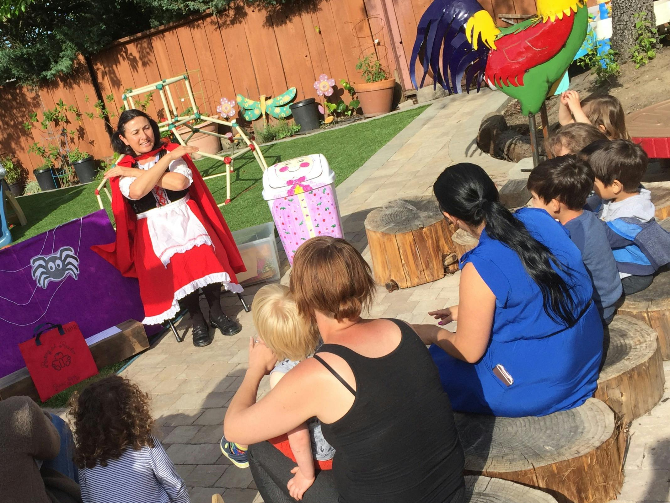 Person in red cape and white apron speaking to adults and children outdoors