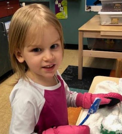 Child wearing pink mittens and holding a blue brush over white material