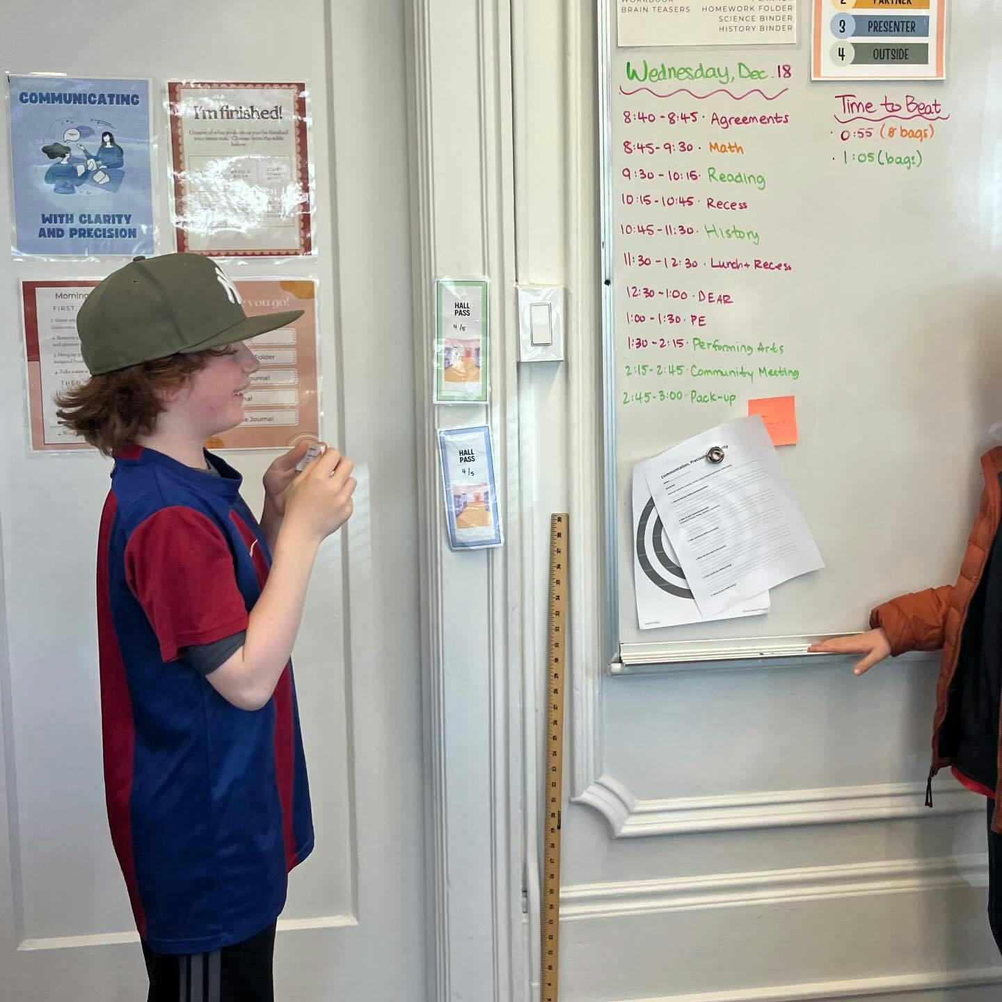 Child wearing a cap and holding a small item next to a whiteboard with a schedule