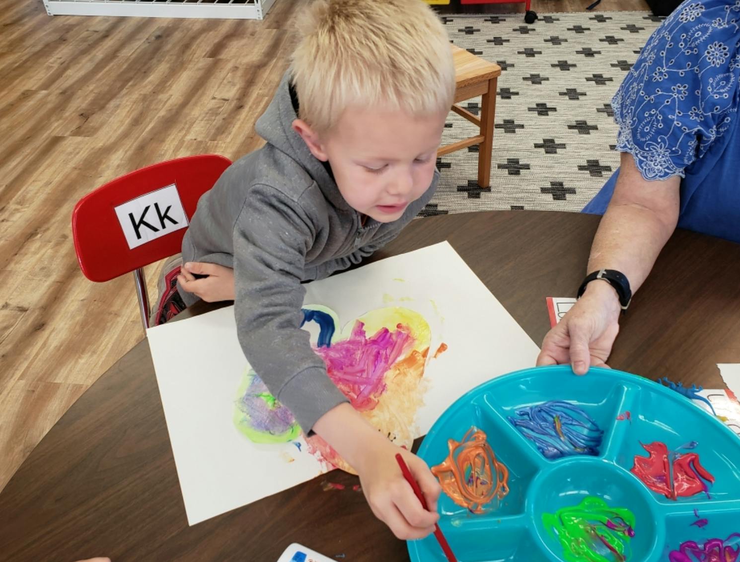 Child painting on paper with a person in a classroom