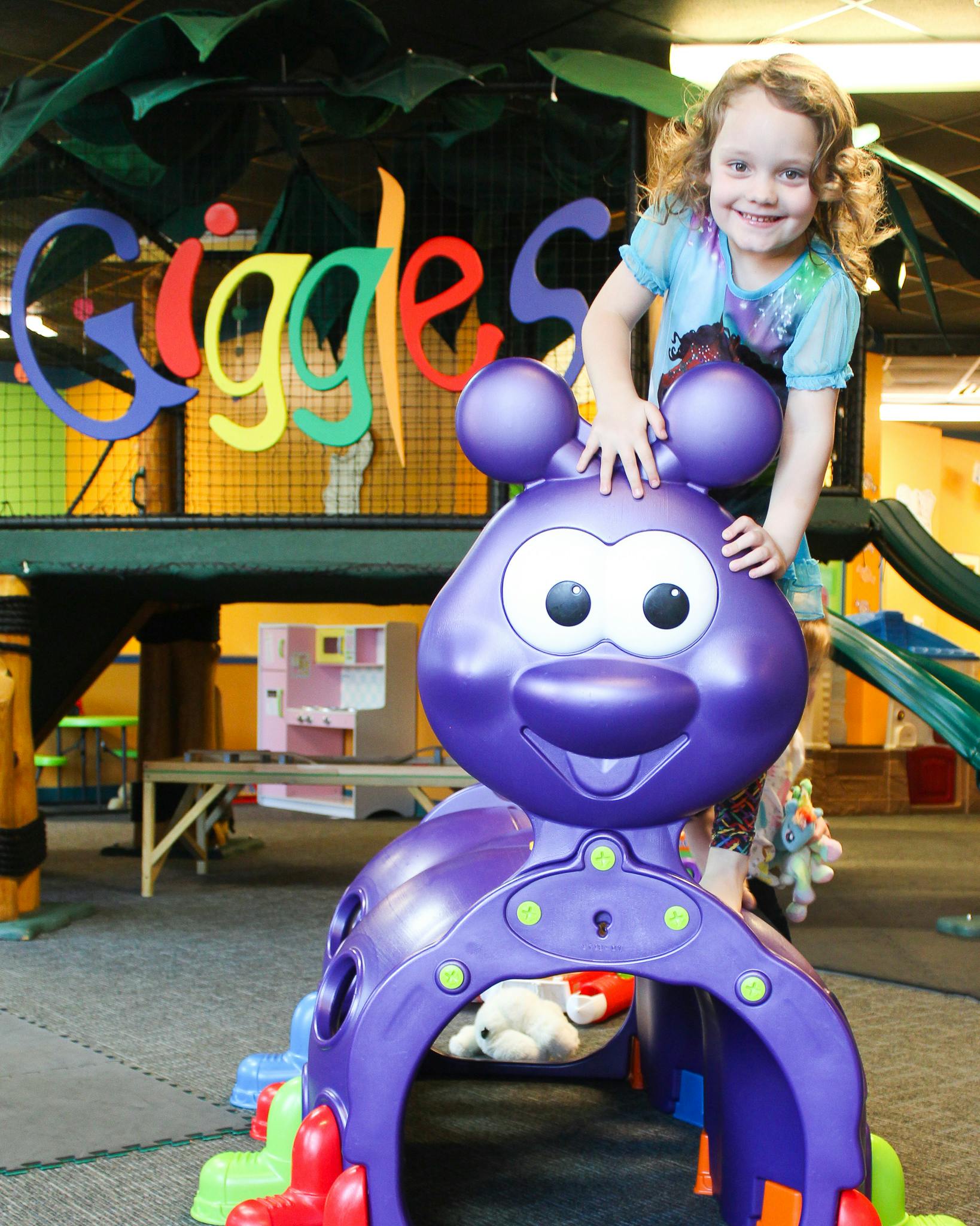 Child standing behind a large purple toy with the word "Giggles" in the background