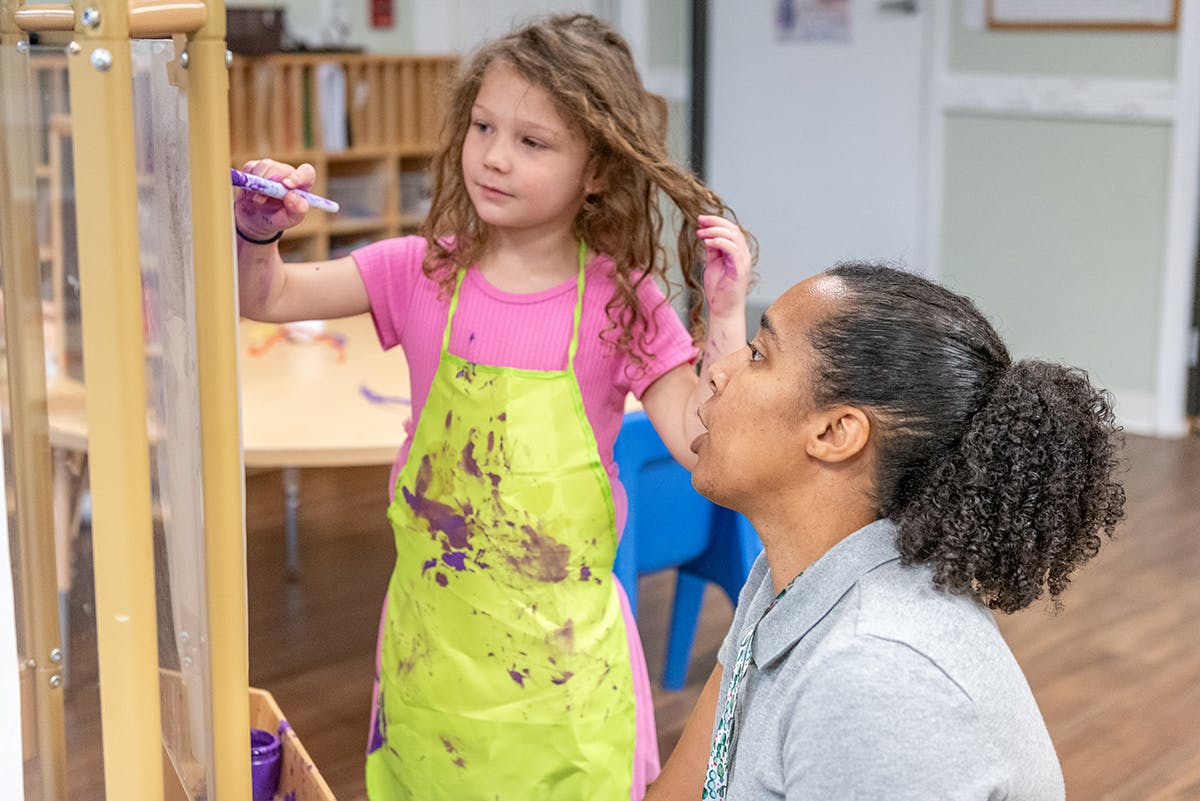 Child in apron painting on easel with adult watching