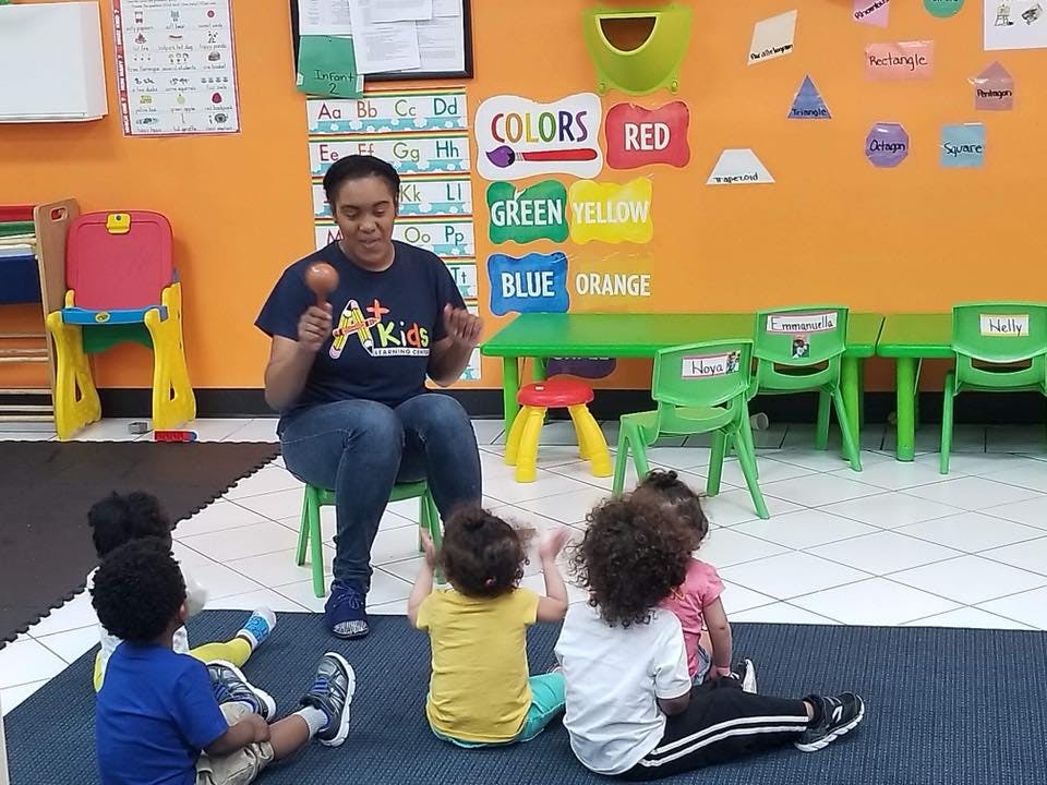 Person holding a maraca, sitting on a chair with children on the floor