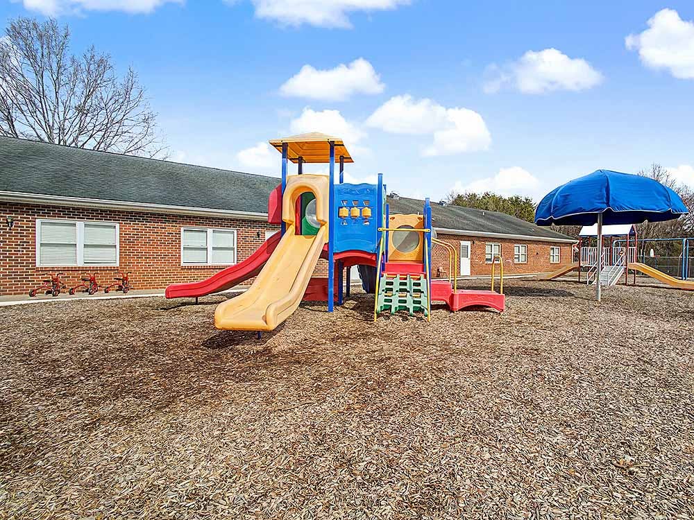 Colorful playground structure with slides on wood chip ground next to a brick building