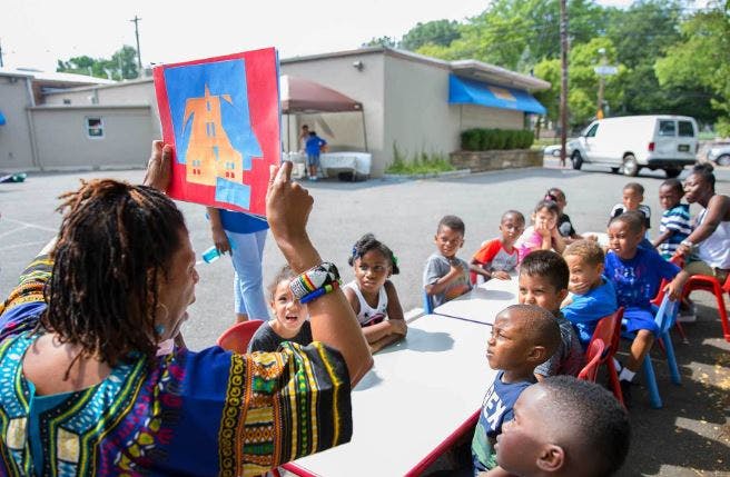 Person holding up a colorful drawing to a group of children seated at tables outdoors