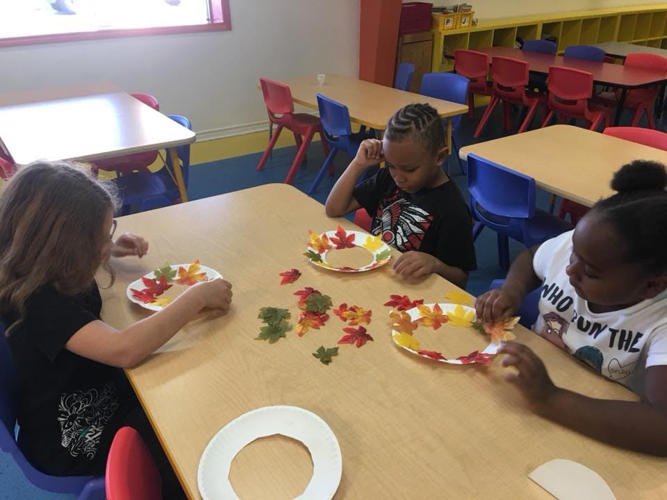 Three children making crafts with paper plates and leaf cutouts in a classroom