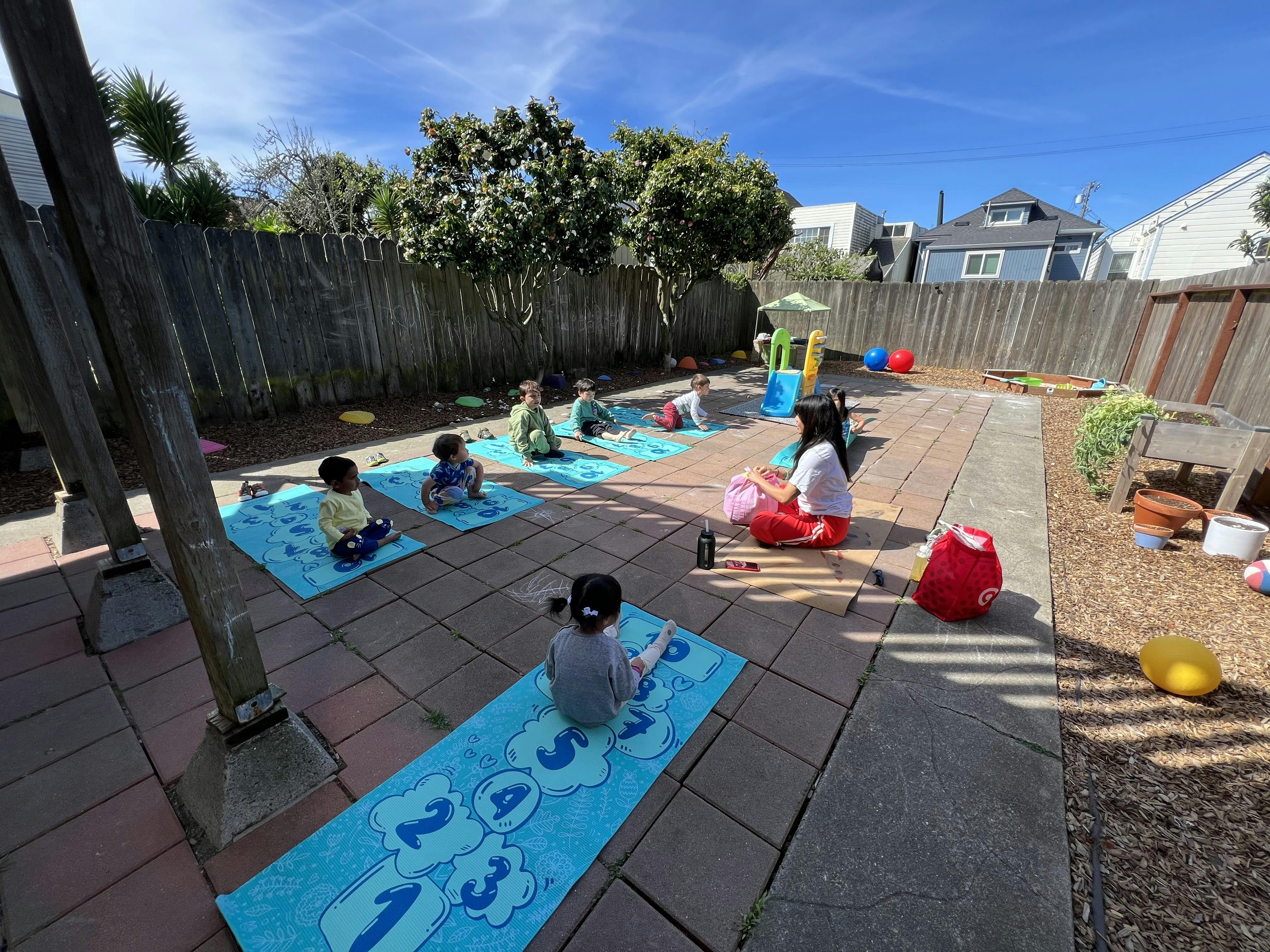 A person leads a group of children in an outdoor yoga session on blue mats
