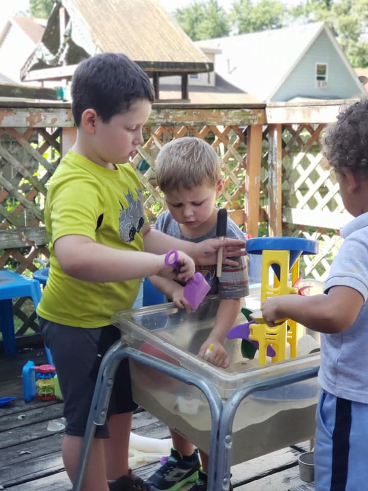 Three children playing with sand and toys in a sensory table outdoors