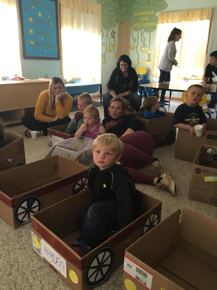 Children and adults sitting on the floor with cardboard box cars