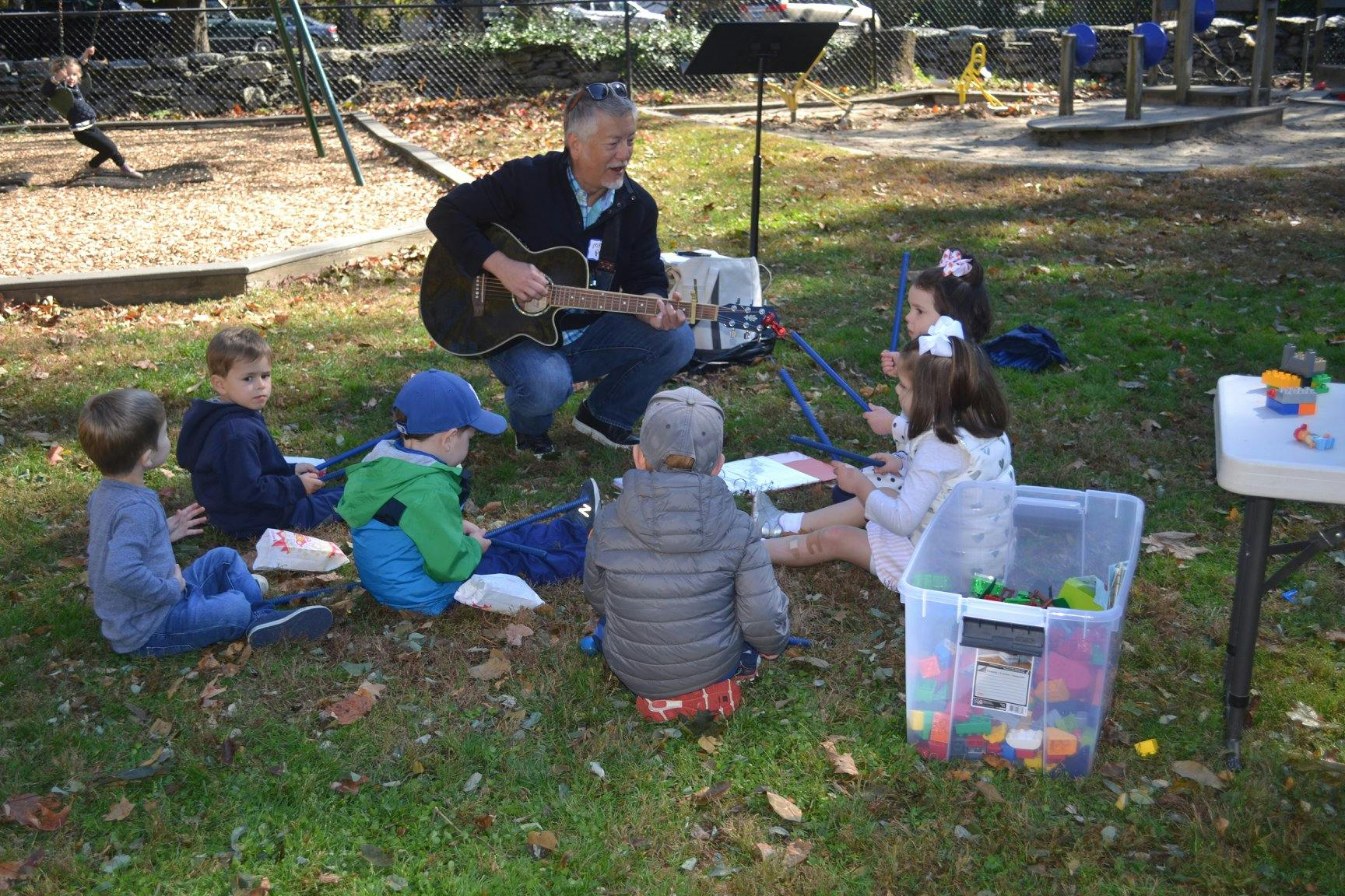 Person playing guitar for a group of children sitting on grass