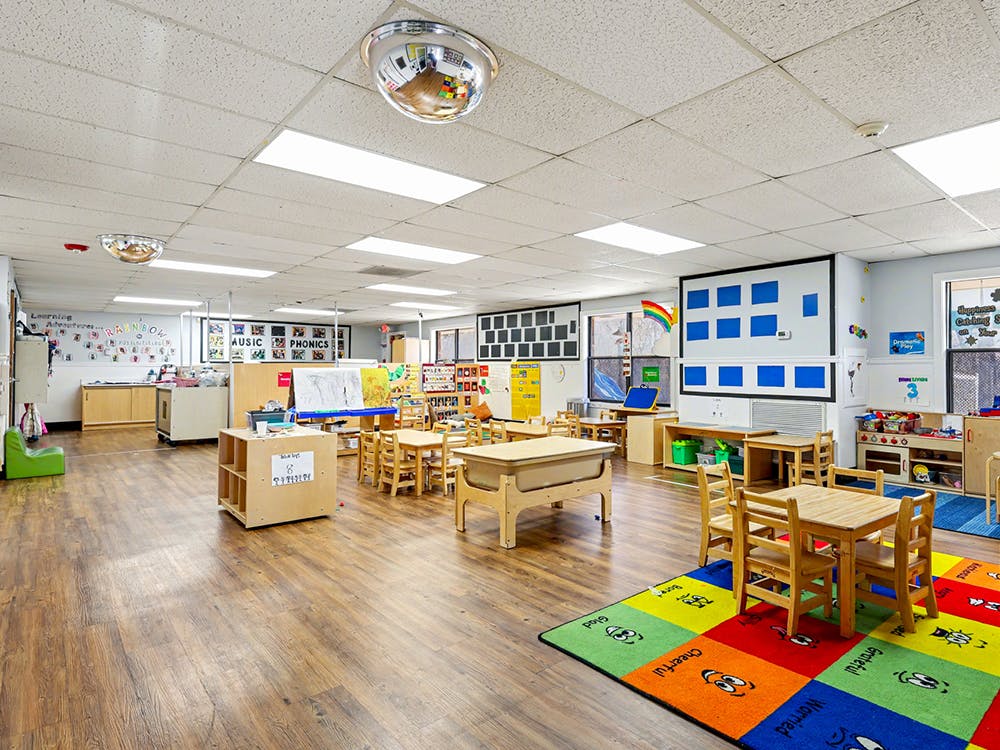 Empty classroom with small wooden tables and chairs, colorful rug, and learning materials