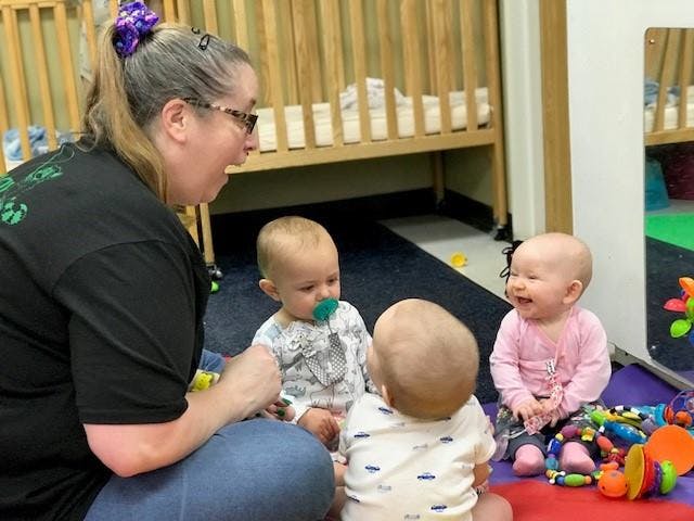 Person interacting with three babies on the floor in a room with cribs in the background