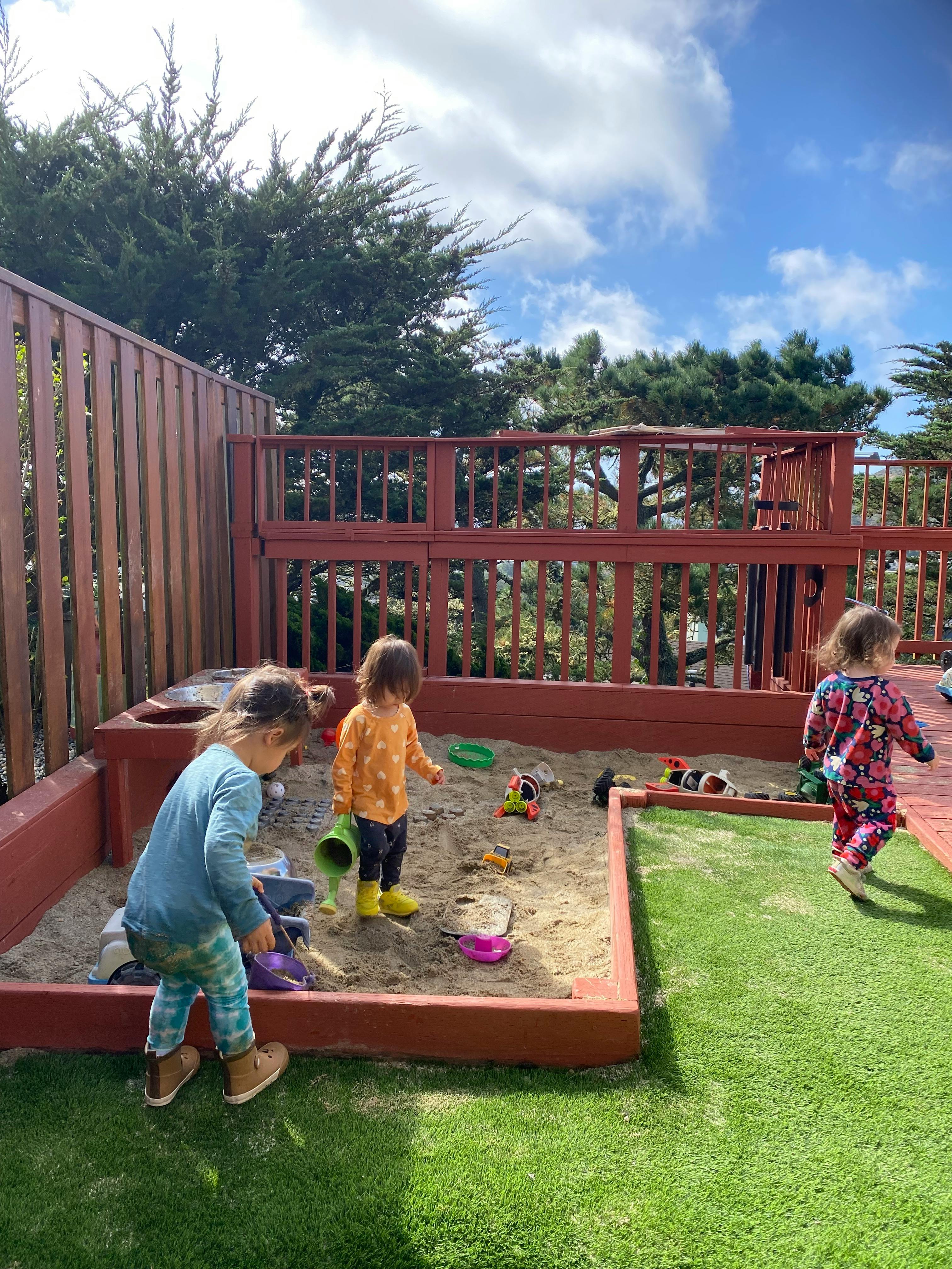 Three children playing in a sandbox and on artificial grass