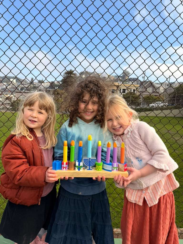 Three children holding a wooden toy menorah with colorful candles