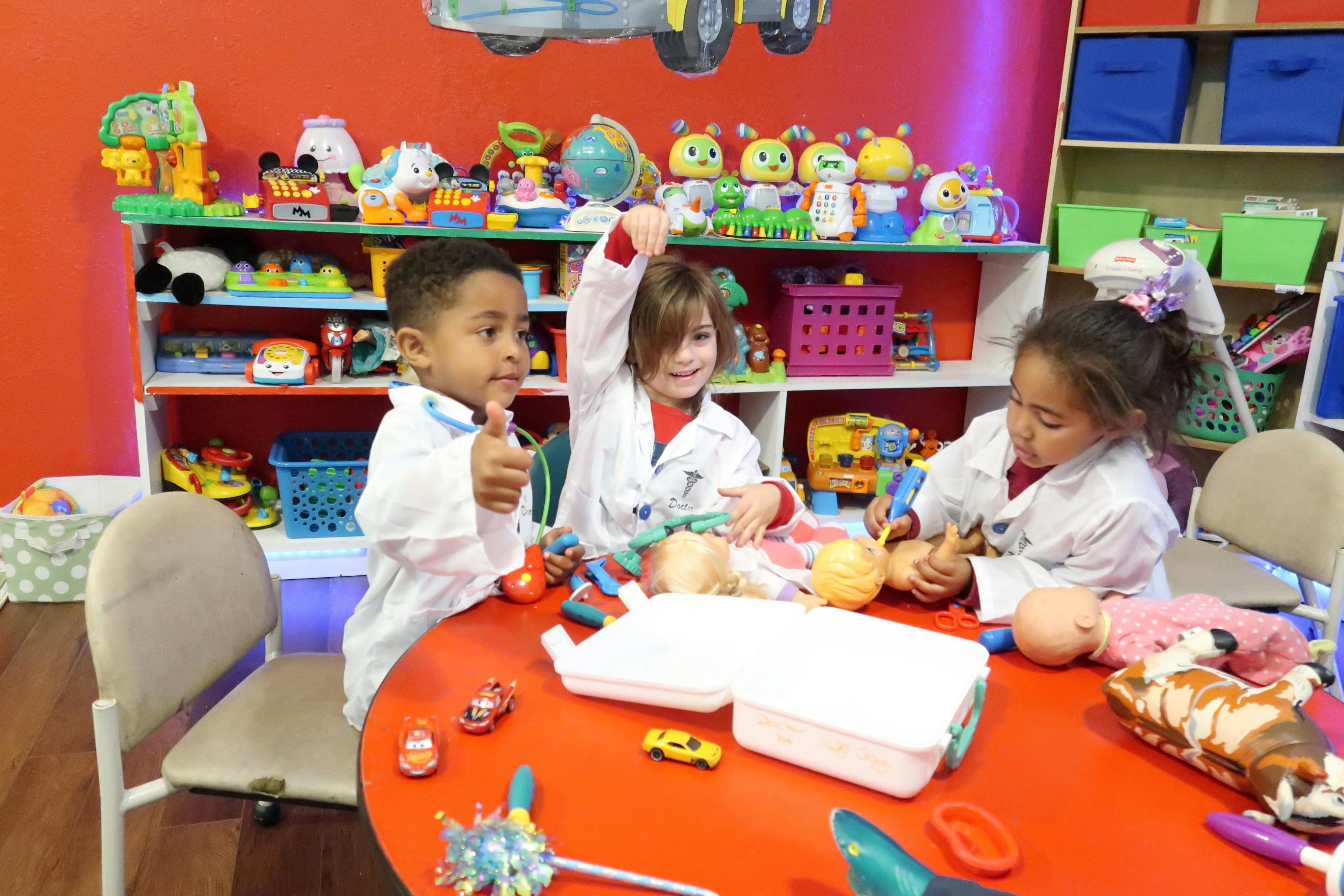 Three children in lab coats playing with toys at a red table