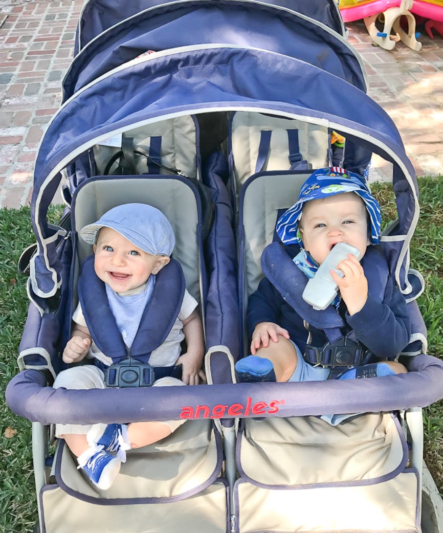 Two children in a double stroller, one smiling and one holding a bottle