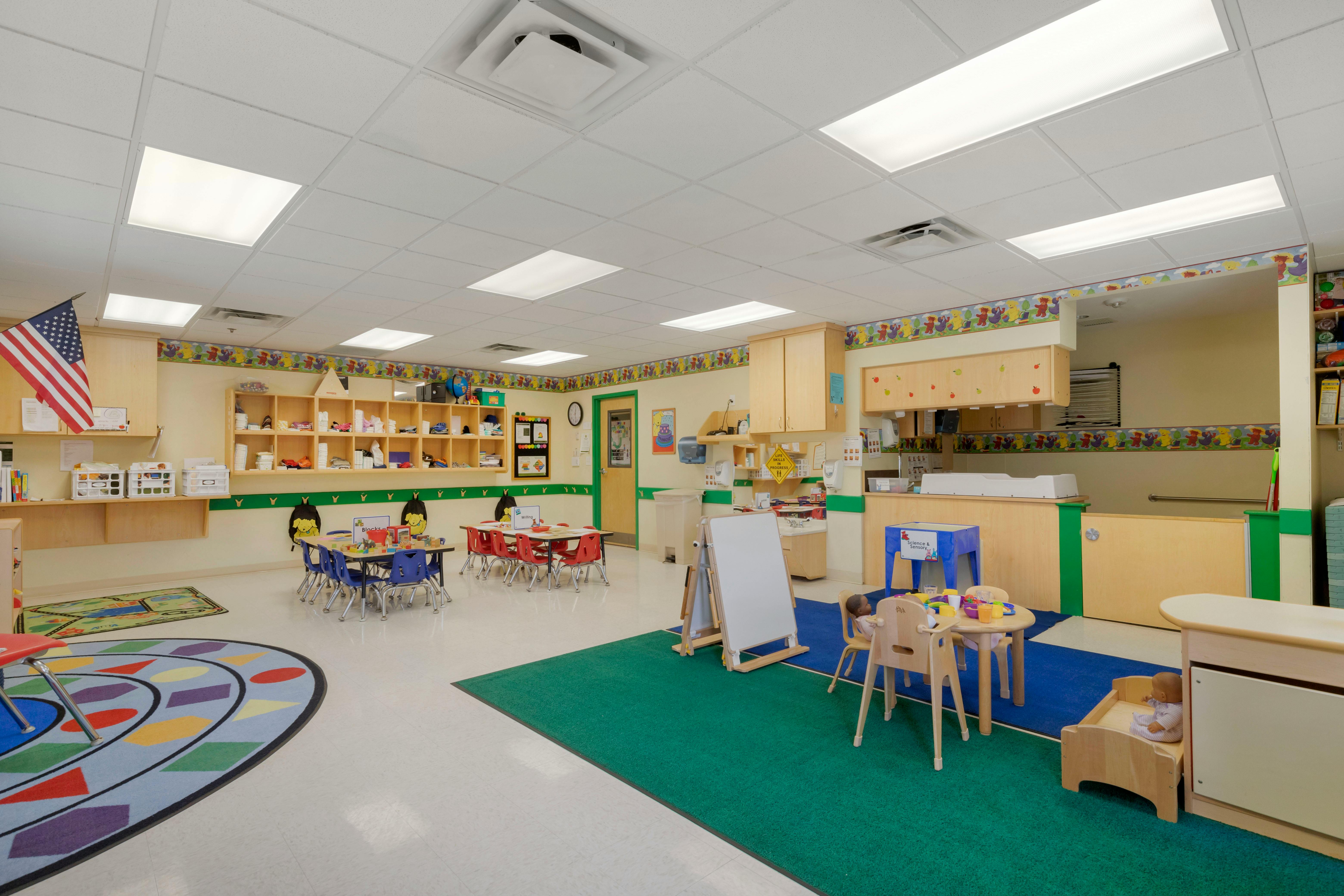A colorful classroom with child-sized tables and chairs, shelves, and rugs