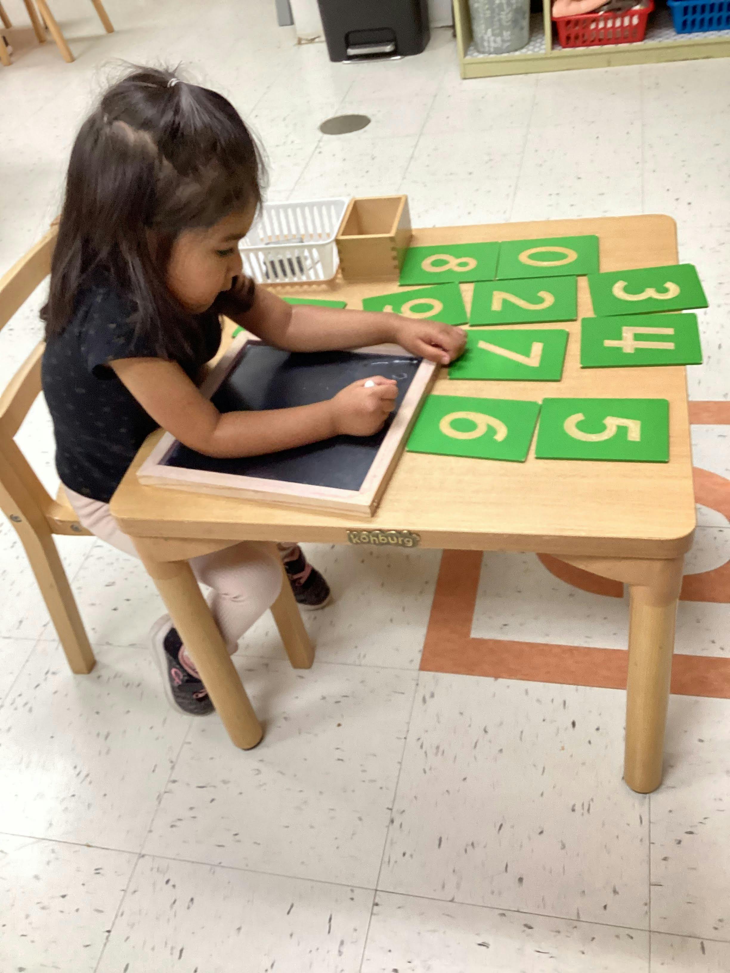 Child writing on a chalkboard at a table with number cards