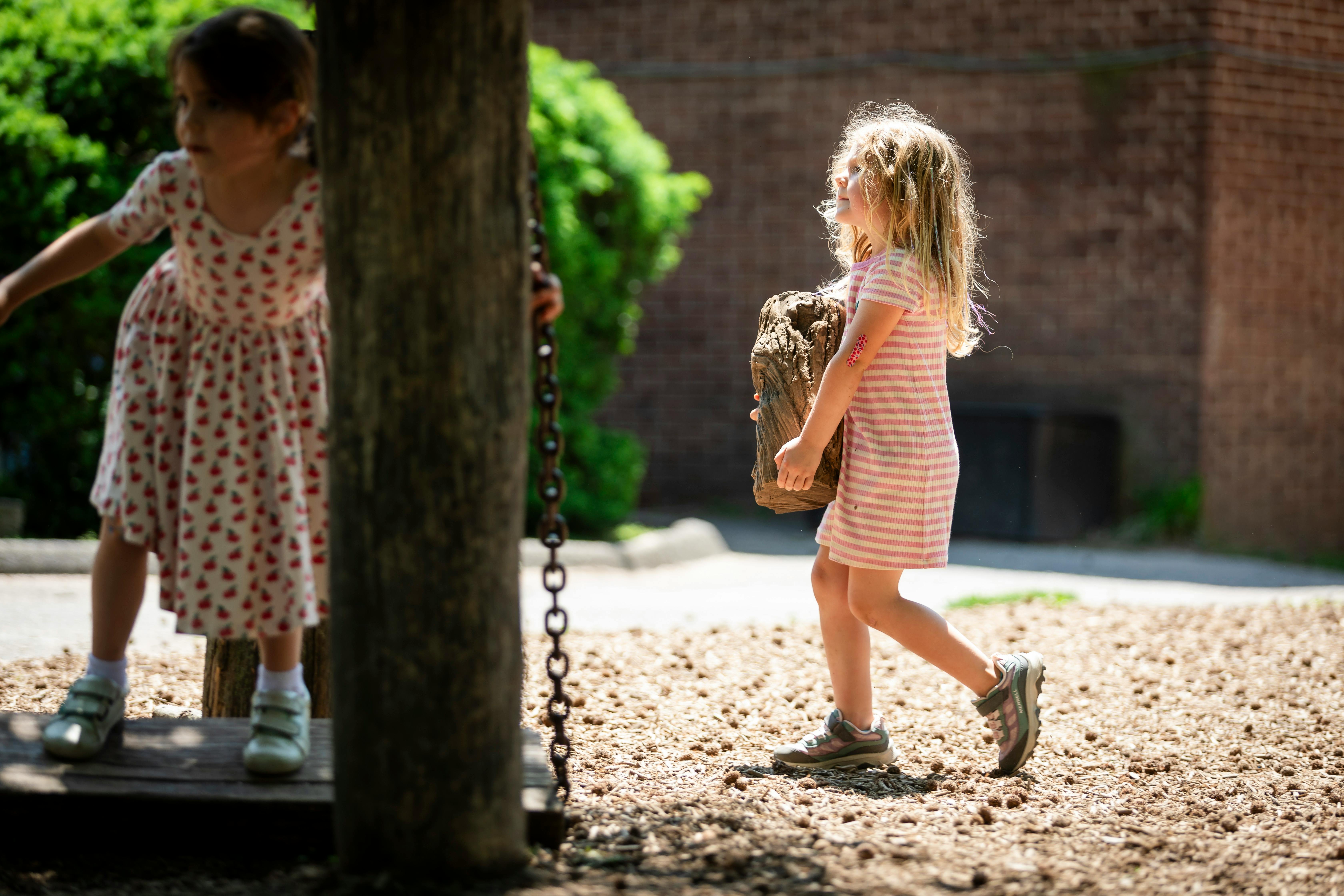 Child carrying a large rock, another child standing behind a tree