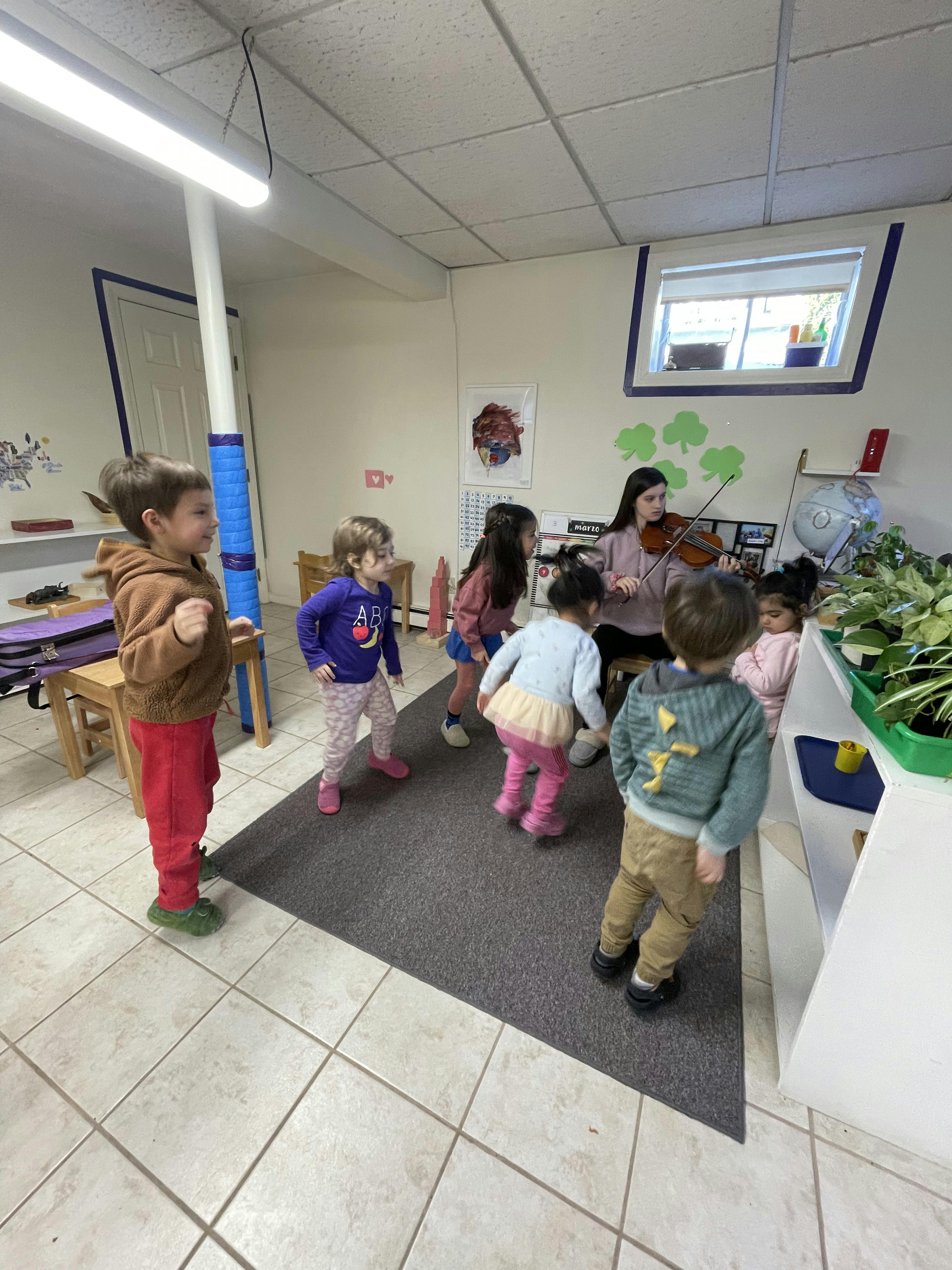 Group of children dancing while an adult plays the violin in a classroom
