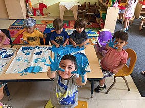 Child with blue paint on hands and face, with other children painting