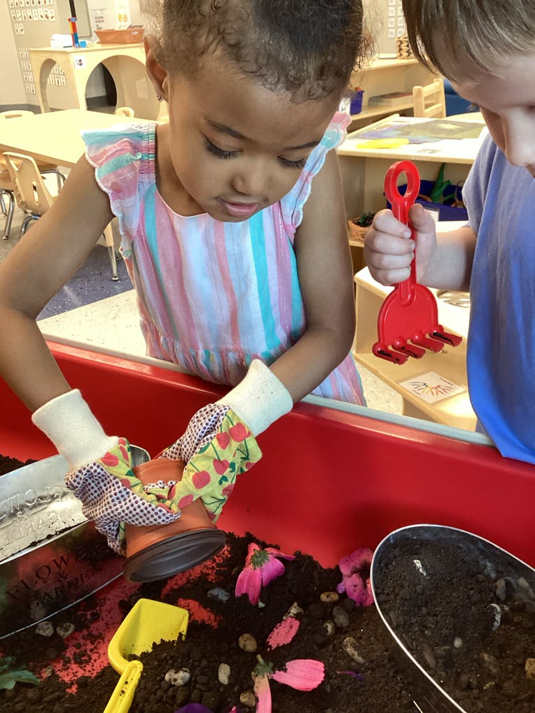 Child wearing gloves planting in a red bin, another child holding a red rake