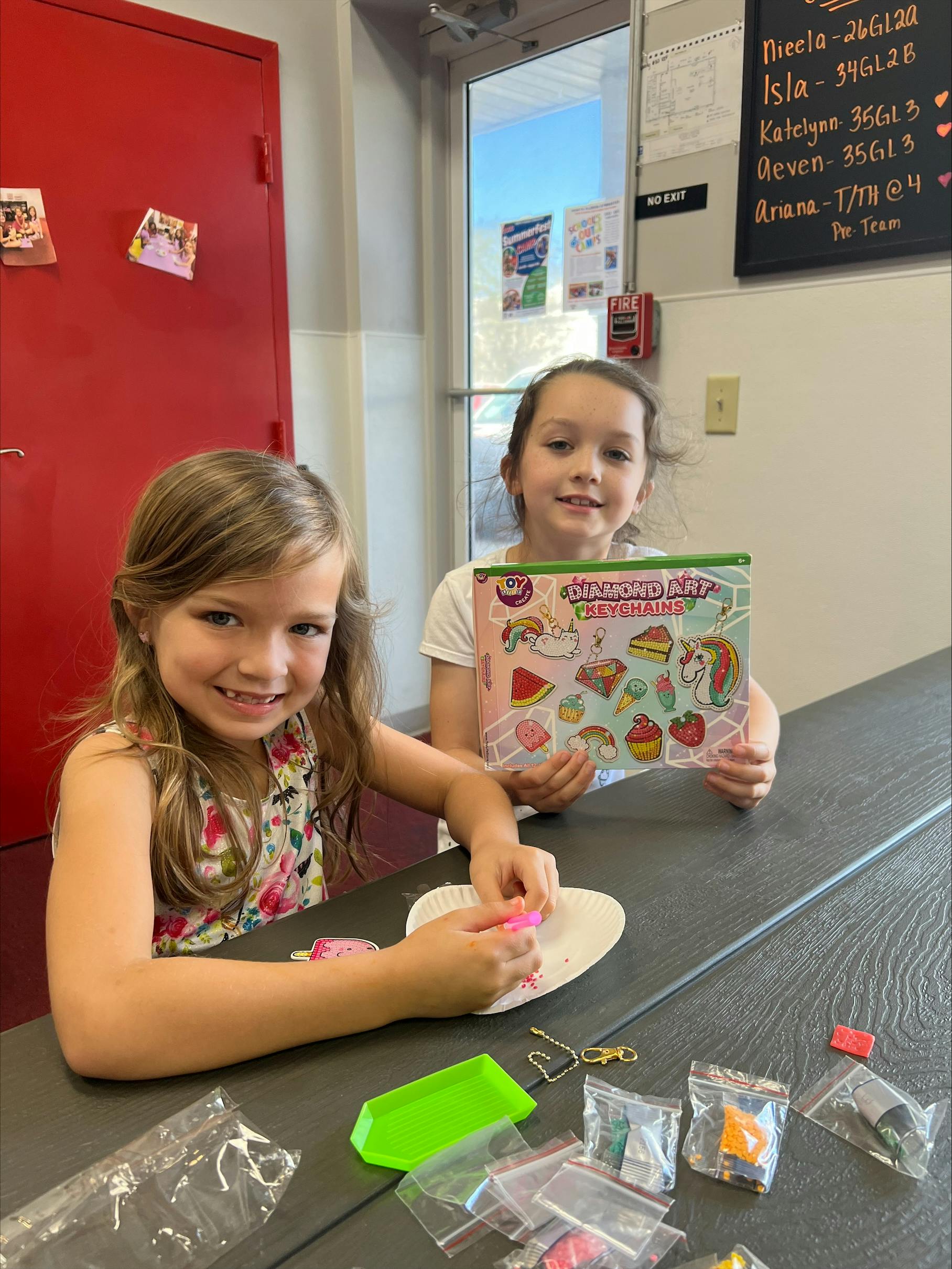 Two children at a table, one holding a craft kit, the other working on a craft