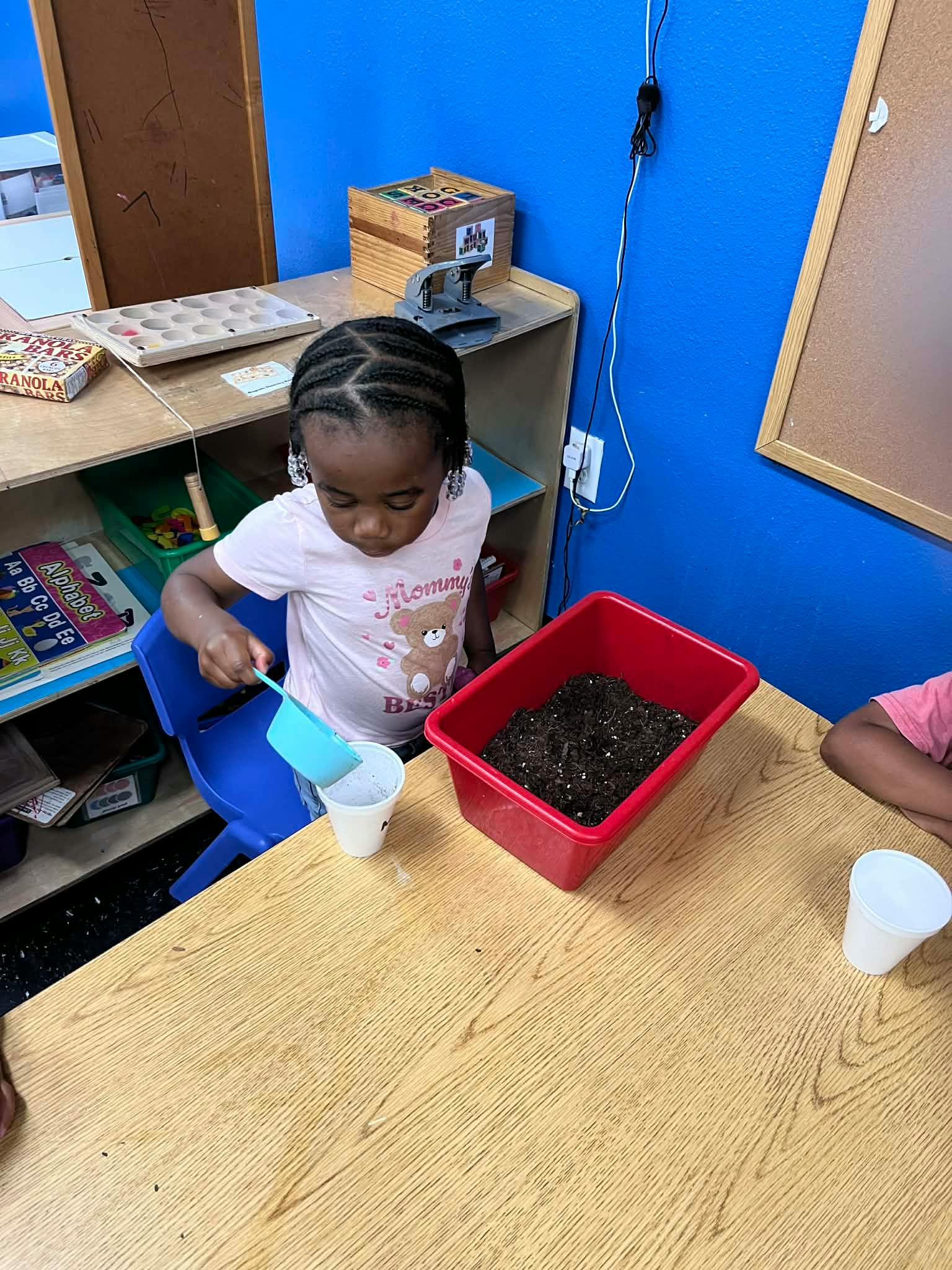 Child pouring dirt from a large red tray into a cup