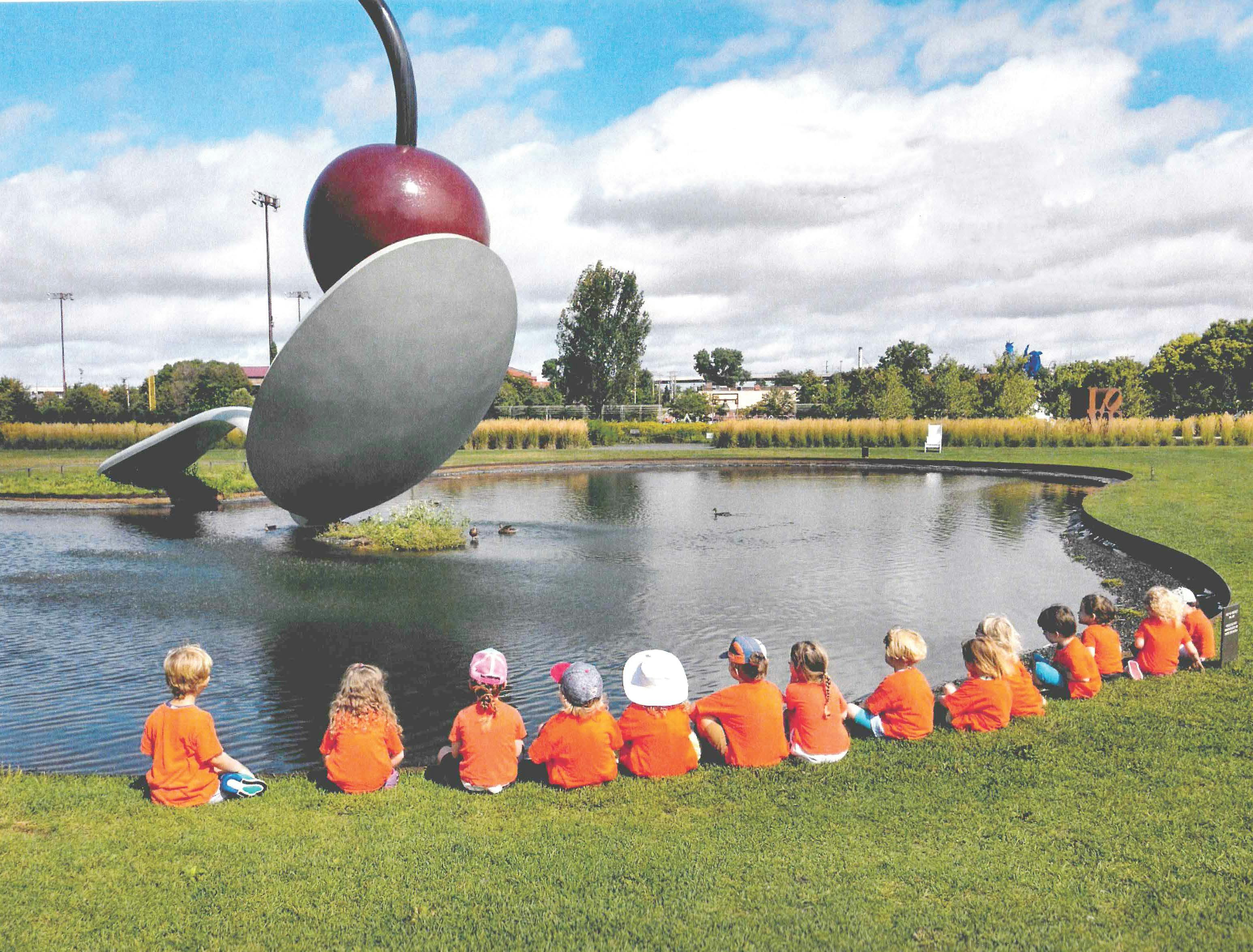 Group of children sitting on grass by a pond facing a large cherry spoon statue