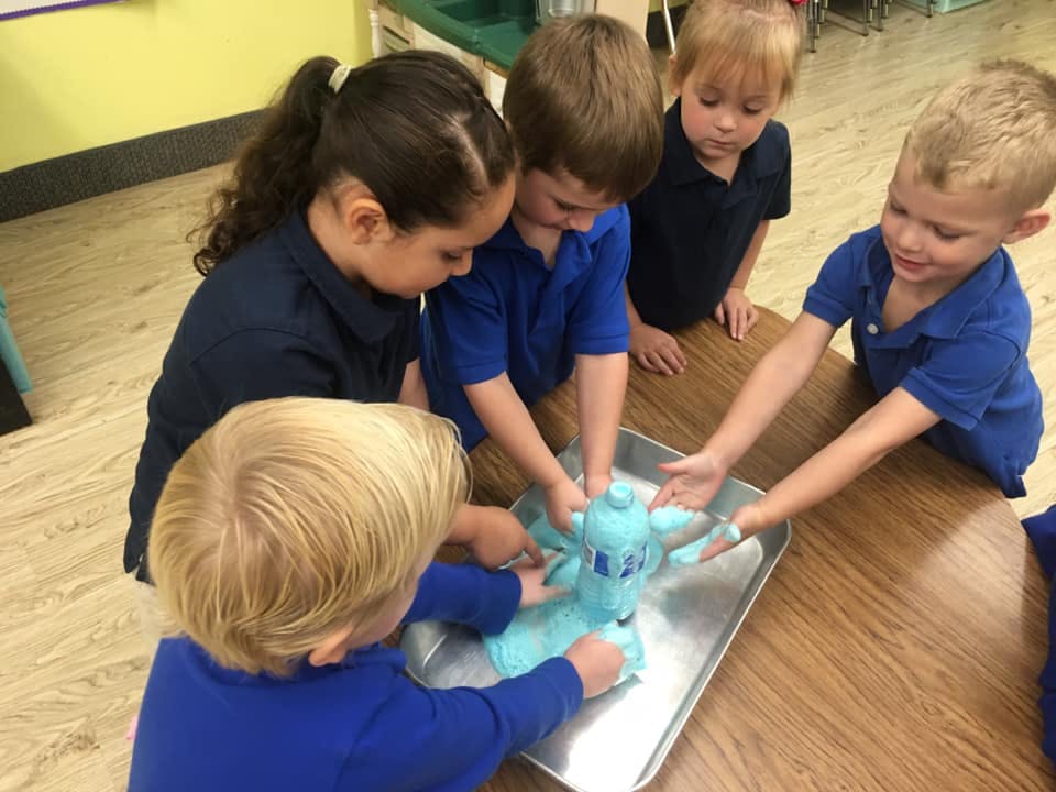 Children gathered around a table, touching a light blue object in a tray