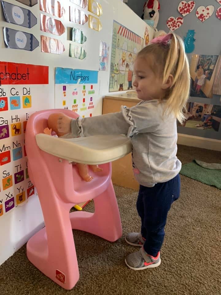 Child standing next to a pink high chair with a doll