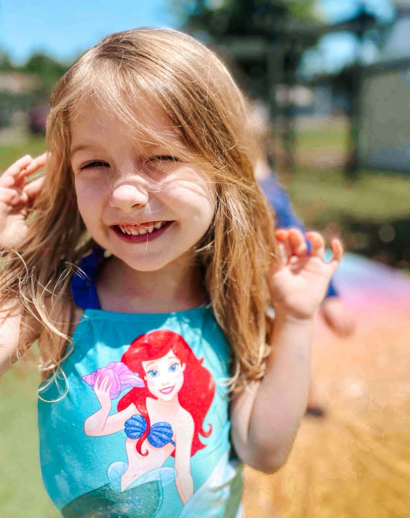 Child smiling outdoors, wearing a shirt with a cartoon mermaid