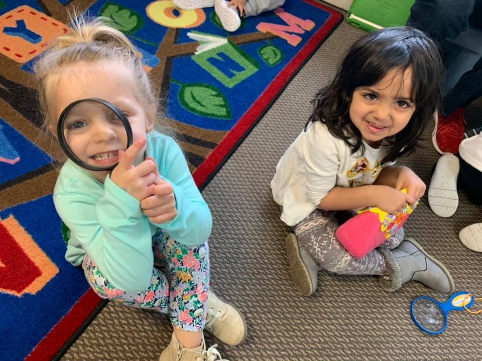 Two children sitting on a rug, one holding a magnifying glass to their eye