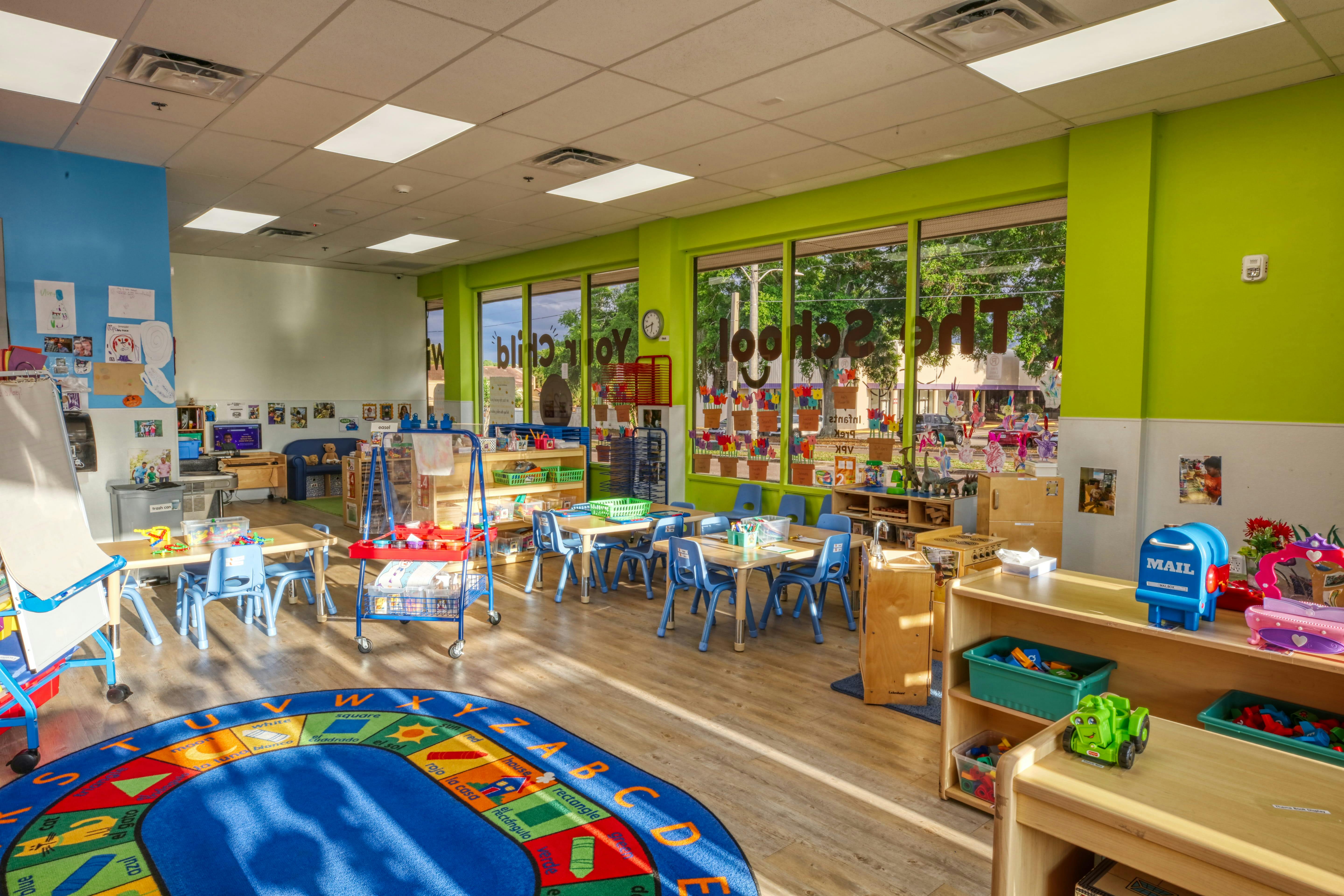 Colorful preschool classroom with tables and chairs, a play rug, and large windows